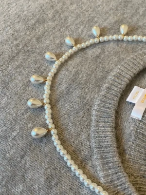 Pärlhalsband med droppformade hängen - Säljer ett elegant halsband med små vita pärlor och droppformade pärlhängen. Halsbandet har guldfärgade detaljer mellan pärlorna och ger en klassisk men trendig look. Perfekt för dig som vill addera något extra till din outfit.