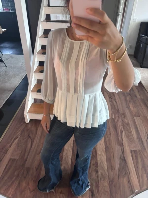 Vit blus med plisserad nederdel - Säljer en vit blus med trekvartsärmar och rund halsringning. Blusen har plisserade detaljer framtill och en volangliknande, plisserad nederdel. Materialet är tunt och lätt transparent, vilket ger en luftig känsla. Perfekt för dig som gillar en romantisk och trendig stil.