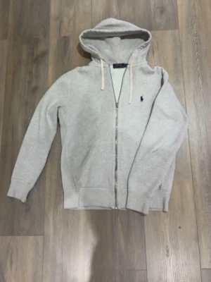 Ljusgrå zip hoodie från Polo Ralph Lauren - Ljusgrå hoodie med dragkedja från Polo Ralph Lauren. Klassisk design med huva och vita dragsnören, samt den ikoniska lilla loggan broderad på bröstet. Tillverkad i mjukt sweatshirtmaterial och har ribbade muddar vid ärmar och nederkant. Perfekt för en avslappnad stil.