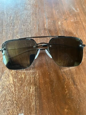 Svarta solglasögon från Prada Nypris 4000kr - Snygga svarta solglasögon från Prada med fyrkantiga glas och metallbågar. Glasen är mörka och bågarna har en tunn, stilren design med diskret Prada-logga på sidan. Perfekta för att höja din look under soliga dagar.