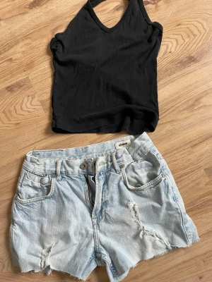 Ljusblå denimshorts från Réunion - Snygga ljusblå denimshorts från Réunion med slitna detaljer och klassisk femficksdesign. Perfekta för varma dagar och en avslappnad stil. Shortsen har bälteshällor och knappgylf.  Halterneck Linne  i svart 