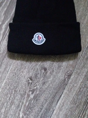 Svart Moncler mössa med logga - Snygg svart ribbstickad mössa från Moncler med uppvikt kant och klassisk Moncler-logga framtill. Perfekt accessoar för att hålla värmen och samtidigt se trendig ut. Materialet är mjukt och stretchigt för skön passform.