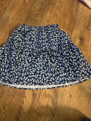 Blommig blå kortkjol med volang - Söt kortkjol i mörkblått med vitt blommönster och volangdetaljer. Kjolen har resår i midjan för skön passform och är perfekt för vår och sommar. Materialet känns lätt och luftigt.
