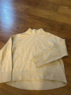 Beige stickad tröja från Kappahl - Mysig beige stickad tröja från Kappahl med hög hals och ribbade muddar vid ärmslut och nederkant. Tröjan har en avslappnad passform och är något längre bak. Perfekt för lager på lager under kyliga dagar. Lite knottrig annars fin