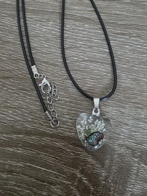 Halsband med hjärthänge och blommor - Unikt halsband med svart läderrem och ett hjärtformat hänge i transparent glas. Hänget innehåller små vita torkade blommor och en fjäril, vilket ger en naturinspirerad och bohemisk känsla. Perfekt accessoar för dig som gillar detaljer och naturmotiv.