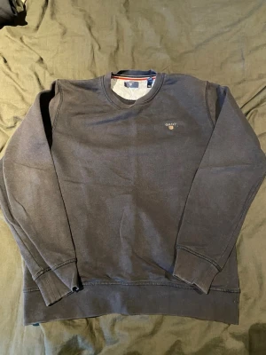 Mörkblå sweatshirt från Gant - Mörkblå sweatshirt från Gant med rund halsringning och diskret logga broderad på bröstet. Tröjan har ribbade muddar vid ärmslut och nederkant samt klassisk passform. Perfekt för en avslappnad och stilren look har även ett litet hål på framsidan.