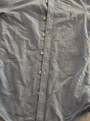 Blå Ralph Lauren skjorta  - Klassisk blå skjorta i bomull med knappar framtill och en broderad logga på bröstet. Skjortan har lång ärm och en tidlös design som passar till många olika stilar.