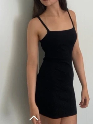 Svart ribbad bodycon klänning - En svart, tight bodycon klänning med ribbat tyg och smala axelband. Klänningen har en rak urringning och kort längd, perfekt för en trendig och enkel look.