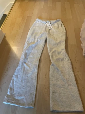 Ljusgrå mjukisbyxor med utsvängda ben - Snygga ljusgrå mjukisbyxor med utsvängda ben och dragsko i midjan. Byxorna har fickor och en broderad detalj på ena fickan. Perfekta för en chill och avslappnad stil. Tillverkade i mjukt och bekvämt material.