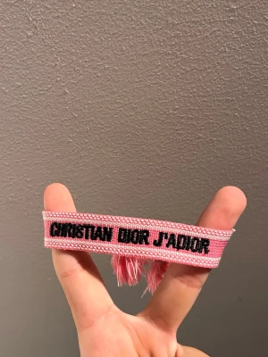 Christian Dior rosa tygarmband - Snyggt rosa armband från Christian Dior med texten 'J'ADIOR' i svart. Armbandet är gjort i vävt tyg och har tofsar i ändarna för en trendig look. Perfekt accessoar för att lyfta din outfit med en lyxig touch.