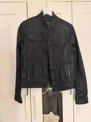 Svart skinnjacka med dragkedjor - Snygg svart skinnjacka med ståkrage och dekorativa bröstfickor med lock. Jackan har dragkedjor vid ärmsluten och framtill, samt detaljerade sömmar för en cool look. Passar perfekt till jeans och sneakers för en trendig outfit. Står inte storlek men skulle säga S/M. 