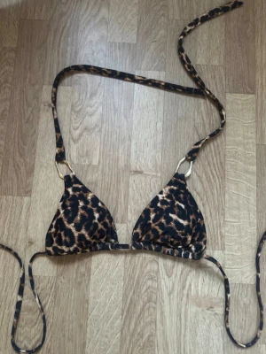 Leopardmönstrad trekantsbikini - Snygg trekantsbikini med leopardmönster och tunna knytband. Bikinin har guldfärgade ringdetaljer vid axelbanden och är fodrad med svart tyg på insidan. Perfekt för strandhäng och poolparty.