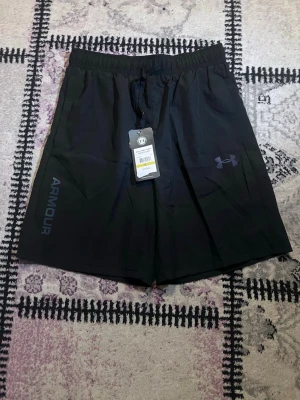 Svarta träningsshorts från Under Armour - Säljer ett par svarta träningsshorts från Under Armour med elastisk midja och snörning. Shortsen har logga på vänster ben och texten 'ARMOUR' på sidan. Tillverkade i ett lätt och snabbtorkande syntetmaterial, perfekta för gym eller löpning.