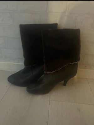 Bruna stövlar med pälsdetalj - Snygga bruna stövlar med hög klack och bred pälsdetalj upptill. Dekorativ rad med små nitar längs kanten mellan skinn och päls. Perfekta för att lyfta din höst- eller vinterstil. Klassisk spetsig tå och elegant siluett.