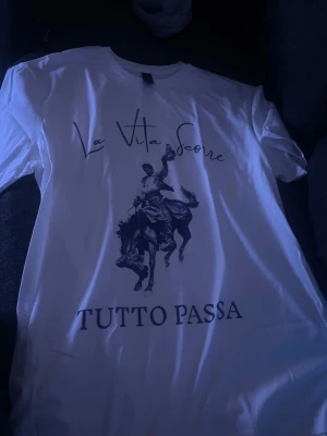 Vit t-shirt med tryck och text - Säljer en vit t-shirt med tryck av en cowboy på häst och texten 'La Vita Scorre' samt 'Tutto Passa' på framsidan. T-shirten har korta ärmar och är i klassisk passform. Perfekt för dig som gillar stilrena plagg med statement-tryck.