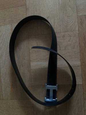 Svart bälte med silverfärgat spänne - Snyggt svart bälte i skinn med ett stort, silverfärgat H-format spänne. Bältet har stilrena sömmar längs kanterna och passar perfekt till jeans eller kostymbyxor för en clean look.