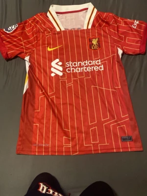 Liverpool matchtröja Szoboszlai #8 - Röd Liverpool matchtröja med gula och vita detaljer, tryckt med Szoboszlai och nummer 8 på ryggen. Tröjan har klubbmärke, Nike-logga och sponsortryck framtill. Tillverkad i lätt och ventilerande material, perfekt för fotboll eller träning.