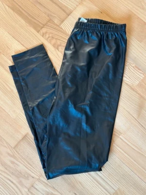Svarta glansiga leggings - Snygga svarta leggings med glansig finish och resår i midjan. De har en tight passform och är perfekta för en trendig look. Materialet ser ut som fuskskinn eller coated tyg som ger en cool vibe.