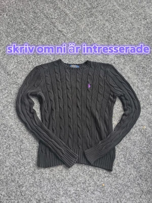Svart stickad äkta Ralph Lauren - Svart kabelstickad tröja från Polo Ralph Lauren med lila broderad logga på bröstet. Tröjan har rund halsringning, långa ärmar och ribbstickade muddar. Perfekt för dig som gillar klassisk och stilren design.