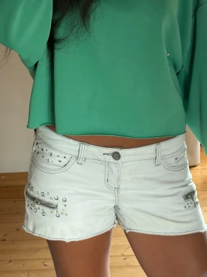 Coola vita lågmidjade jeansshorts  - Snygga vita denimshorts från Melrose i storlek 38 med unika detaljer! Midjemåttet är 39cm. Modellen har en rå, fransig kant nertill och är dekorerad med dekorativa dragkedjor samt glittriga nitar och strassdetaljer både fram och på ena bakfickan. Perfekta till sommarens alla outfits och festivaler. I fint skick – hör av dig vid frågor! ✨