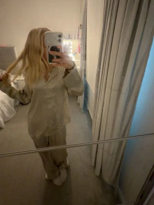 Beige satinpyjamas - Säljer en beige pyjamas i mjukt satinmaterial med långärmad skjorta och matchande byxor. Perfekt för mysiga kvällar hemma. Säljer båda delarna ihop!