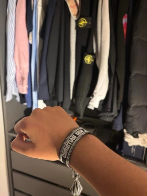 Dior armband - Snyggt armband från Christian Dior med texten 'J'Adior' i svart och vitt. Armbandet är vävt i tyg och har dekorativa tofsar i ändarna. Perfekt accessoar för att lyfta din outfit med en exklusiv touch.