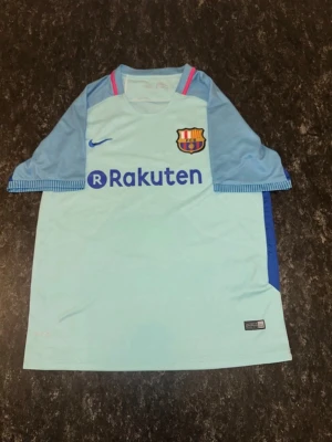 Ljusblå FC Barcelona matchtröja - Säljer en ljusblå FC Barcelona matchtröja från Nike med klubbmärke och Rakuten-logga på bröstet. Tröjan har blå detaljer på ärmarna och rosa inslag vid kragen. Tillverkad i ett lätt och ventilerande material, perfekt för fotbollsträning eller match.
