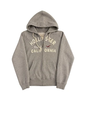 Grå hoodie från Hollister med dragkedja - Säljer en grå hoodie från Hollister med dragkedja och broderad text 'Hollister California 1922' på bröstet. Hoodien har vita snören, två fickor fram och mjukt, fluffigt foder på insidan. Perfekt för chill dagar och streetwear vibes.