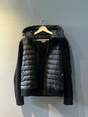 Svart pufferjacka från Moncler - Snygg svart pufferjacka från Moncler med stickade ärmar och vadderad front. Jackan har huva, dragkedja framtill och ribbade muddar. Perfekt för kalla vinterdagar och ger en sportig men stilren look.