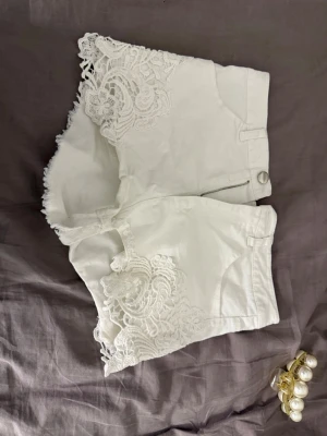 Vita shorts med spetsdetaljer - Snygga vita shorts med spetsdetaljer längs bensluten och råa kanter. De har klassisk femficksdesign, dragkedja och knapp framtill. Perfekta för sommarens alla äventyr och ger en fräsch och trendig look.