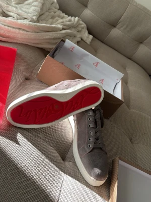 Gråa sneakers med röd sula - Snygga grå sneakers i mocka med klassisk röd sula och vita kanter. Skorna har lågt skaft och matchande grå skosnören. Perfekta för dig som vill ha en stilren och trendig look.