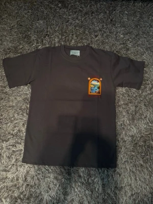 Mörkgrå Casa Blanca t-shirt - Säljer en mörkgrå t-shirt med färgglad tryck fram och bak från Casa Blanca. Motivet visar ett fönster med utsikt över en hamn och blommor. T-shirten har rund hals och korta ärmar, perfekt för dig som gillar unika prints.