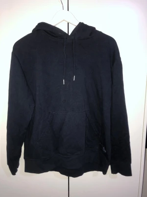 Svart hoodie med magficka och huva - En klassisk svart hoodie med stor magficka och justerbar dragsko i huvan. Hoodien har långa ärmar och ribbade muddar vid ärmslut och nederkant. Perfekt för en avslappnad och chill stil. Materialet ser ut att vara mjukt och bekvämt, troligen bomull eller bomullsblandning.