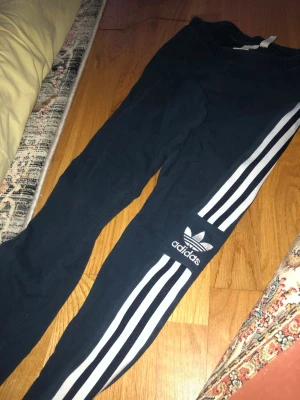 Svarta Adidas mjukisbyxor med ränder - Säljer ett par svarta mjukisbyxor från Adidas med de klassiska vita tre ränderna längs benen och Adidas-logga på låret. Byxorna har smal passform och resår i midjan. Perfekta för chill eller träning.