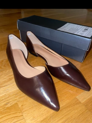 Bruna ballerinaskor i skinn strl 41 - Oanvända/ helt nya- Säljer ett par stilrena ballerinaskor i mörkbrunt skinn med spetsig tå och låg klack. Skorna har en klassisk och elegant design med slät yta och beige innersula. Perfekta för dig som gillar minimalistisk och tidlös stil.(CN43)