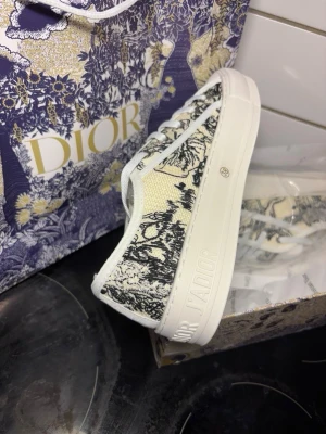 Dior sneakers med svartvitt mönster - Snygga Dior sneakers i lågt snitt med vit sula och svartvitt toile de Jouy-mönster på textilen. Skorna har vita snören och detaljer med Dior-logga på sidan och sulan. Perfekta för dig som vill sticka ut med exklusiv design och coola detaljer.