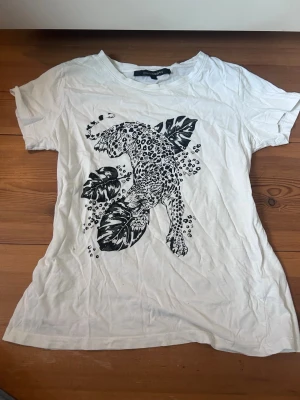 Vit t shirt - Vit t shirt med somrigt svart tryck av dn leopard på framsidan. Inte använd mycket, så väldigt bra skick. Strl S. Skriv om du har frågor! Observera de två pyttesmå hålen på framsidan( syns endast om man letar efter dem)