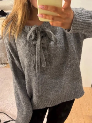 Grå stickad tröja med snörning - Mysig grå stickad tröja med långa ärmar och snörning framtill vid halsringningen. Tröjan har ribbade muddar och en avslappnad passform, perfekt för kyliga dagar. Materialet känns mjukt och behagligt mot huden.