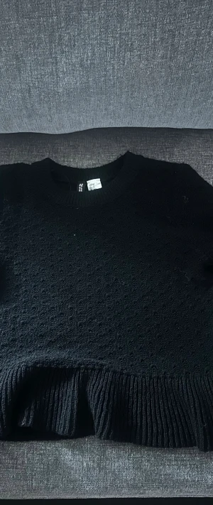 Svart stickad tröja från H&M - Snygg svart stickad tröja från H&M med rund halsringning och diskret strukturmönster. Tröjan har kort ärm och bred ribbad kant nedtill som ger en trendig look. Perfekt att matcha med jeans eller kjol för en cool vardagsstil.
