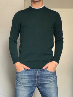 Mörkgrön rundhalsad tröja - En stilren mörkgrön tröja med rund halsringning och långa ärmar. Tröjan har ribbade muddar vid ärmslut och nederkant samt en diskret detalj vid halsen. Perfekt att matcha med jeans för en clean look.
