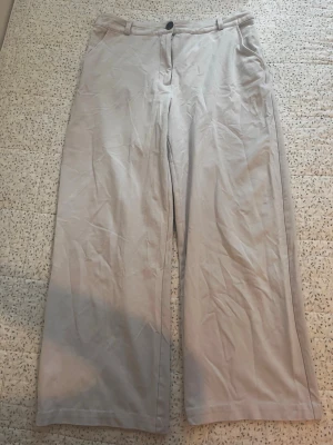 Vida beige chinos med hög midja - Säljer ett par ljusbeige chinos med vida ben och hög midja. Byxorna har knapp och dragkedja framtill samt bälteshällor. Tillverkade i ett mjukt och lätt tyg som passar perfekt till en avslappnad stil.