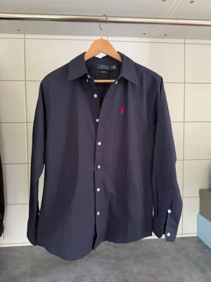 Mörkblå skjorta från Polo Ralph Lauren - Klassisk mörkblå skjorta från Polo Ralph Lauren med vit knappstängning och röd broderad logga på bröstet. Skjortan har lång ärm, normal passform och är tillverkad i bomull. Perfekt för en clean och stilren look.