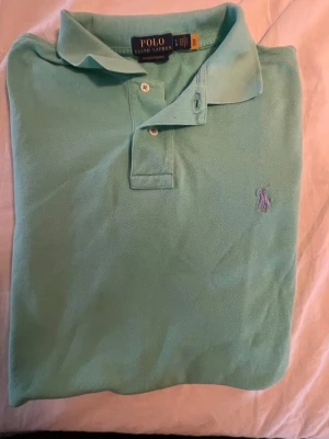 Mintgrön äkta pikétröja från Polo Ralph Lauren - Snygg mintgrön pikétröja från Polo Ralph Lauren med klassisk krage och två knappar framtill. Tröjan har korta ärmar och den ikoniska broderade loggan på bröstet. Perfekt för en avslappnad men stilren look.