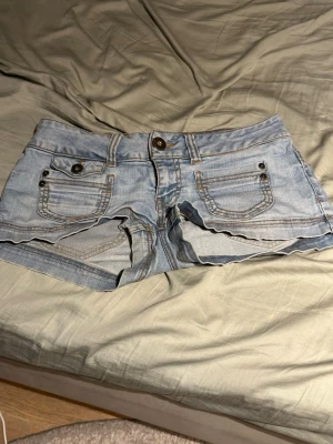 Ljusblå jeansshorts med fickor - Säljer ett par ljusblå jeansshorts med fem fickor, bälteshällor och knappgylf. Klassisk denimlook med kontrastsömmar och nitar på fickorna. Perfekta för varma dagar och enkel att matcha med olika stilar. Liten fläck på vänstra bakfickan men går bort i tvätten.