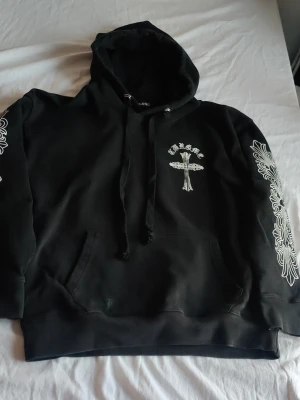 Svart Chrome Hearts hoodie med tryck - Säljer en svart hoodie från Chrome Hearts med vita tryck på bröstet och längs båda ärmarna. Hoodien har en stor huva, dragsko och känguruficka framtill. Materialet är mjukt och skönt, perfekt för en avslappnad streetwear-look.