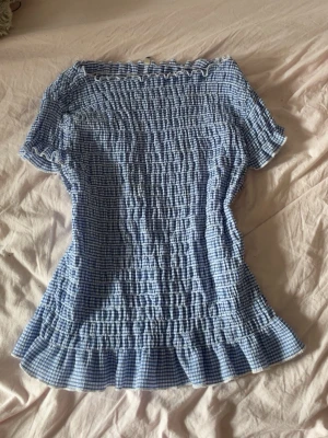 Blårutig smockad topp med volang - Supersöt blårutig topp med smockad struktur och volangkant nedtill. Toppen har korta ärmar och en offshoulder-design som ger en trendig vibe. Perfekt för dig som gillar en feminin och somrig stil.