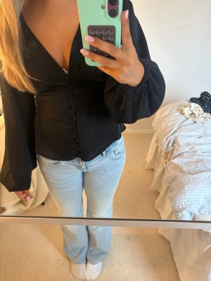 Svart blus med knappar från BYIC - Snygg svart blus från BYIC med v-ringning och dekorativa knappar framtill. Blusen har långa, vida ärmar med resår vid handlederna som ger en trendig look. Tillverkad i ett mjukt, lätt material som sitter fint på kroppen.