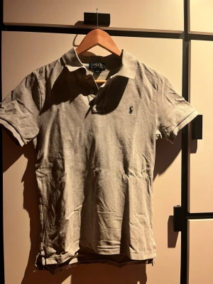 En grå pikétröja från Polo Ralph Lauren - Klassisk grå pikétröja från Polo Ralph Lauren med korta ärmar och krage. Tröjan har den ikoniska broderade loggan på bröstet och är tillverkad i mjuk bomull. Perfekt till jeans eller shorts för en avslappnad men snygg look.
