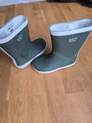 Gröna gummistövlar med vit kant - Säljer ett par klassiska gröna gummistövlar med vit kant upptill och vit sula. Stövlarna har en enkel och stilren design med siffran 157 tryckt på sidan. Perfekta för regniga dagar och leriga äventyr.