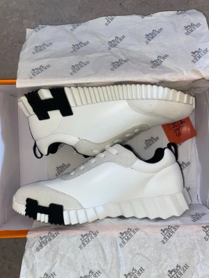 Vita sneakers med taggig sula - Säljer ett par vita sneakers med unik taggig sula och svarta detaljer på sidan. Skorna har en låg profil, vita skosnören och kombinerar slätt läder med mockaliknande partier. Perfekta för dig som vill sticka ut med din stil.
